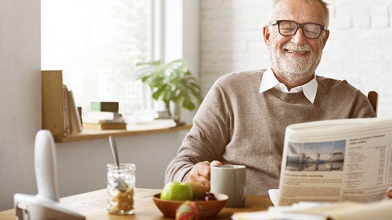 Senior man reading a newspaper while sitting at a kitchen table.