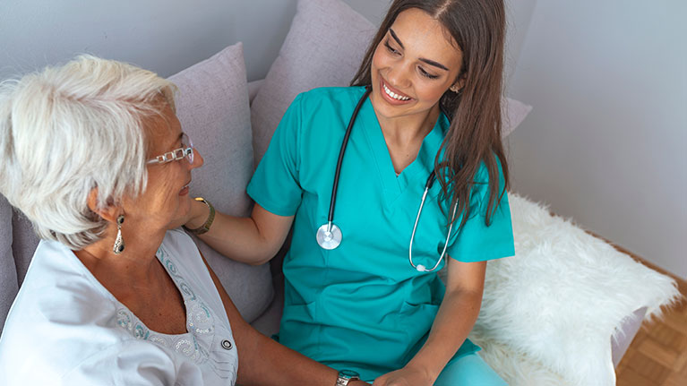 Female nurse in teal scrubs sits on a couch with an elderly female patient smiling.