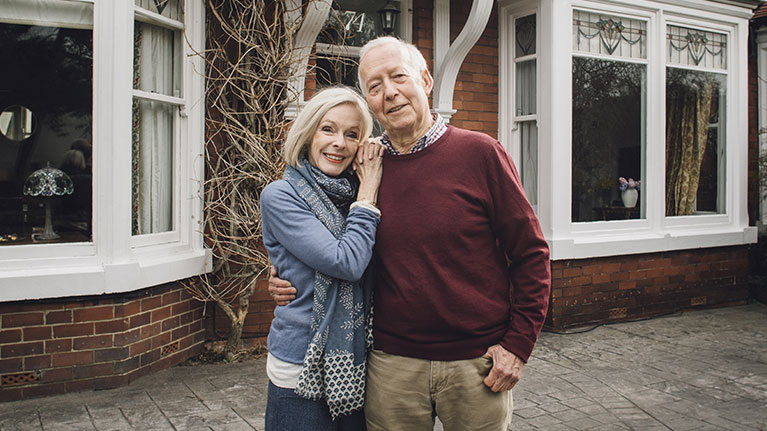 Senior couple embracing and smiling in front of residential building.