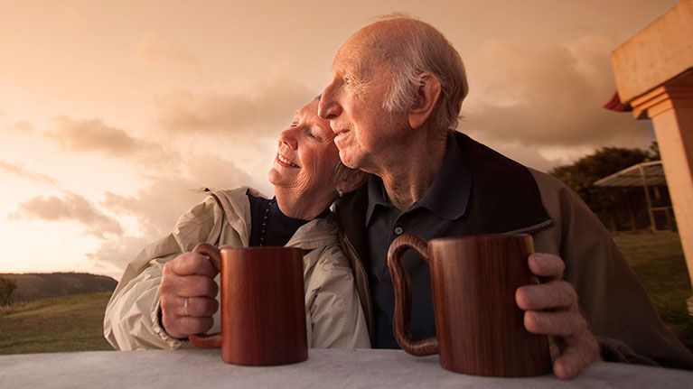 Picture of older couple sitting down holding their coffeemugs while looking out into the field and sunset.