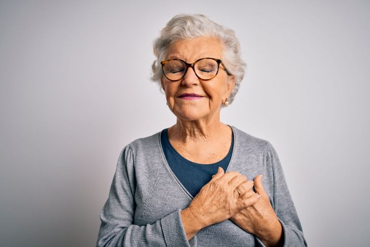 Older woman with glasses holding her hands over her heart.